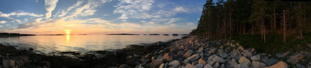 panorama of a sunrise over the ocean, seen from a rocky sea wall adjacent to a forest.